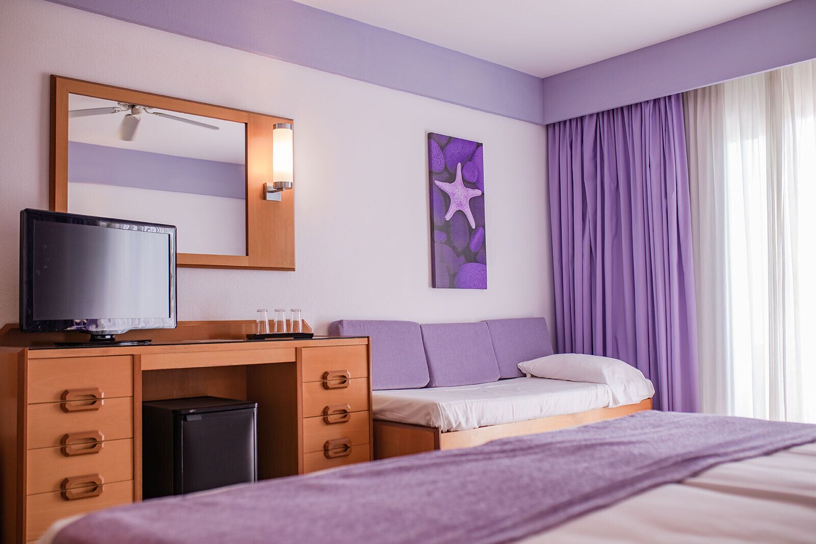 Une chambre d’hôtel avec un lit, un bureau équipé d’un ordinateur et d’une télévision, une petite poubelle, un miroir et une fenêtre avec des rideaux violets.