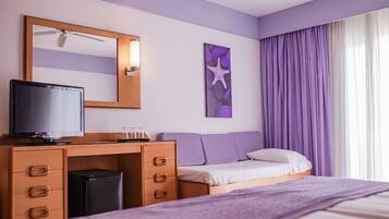 Une chambre d’hôtel avec un lit, un bureau équipé d’un ordinateur et d’une télévision, une petite poubelle, un miroir et une fenêtre avec des rideaux violets.