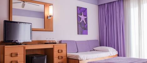 A hotel room with a bed, a desk with a computer and a television, a small trash bin, a mirror, and a window with purple curtains.