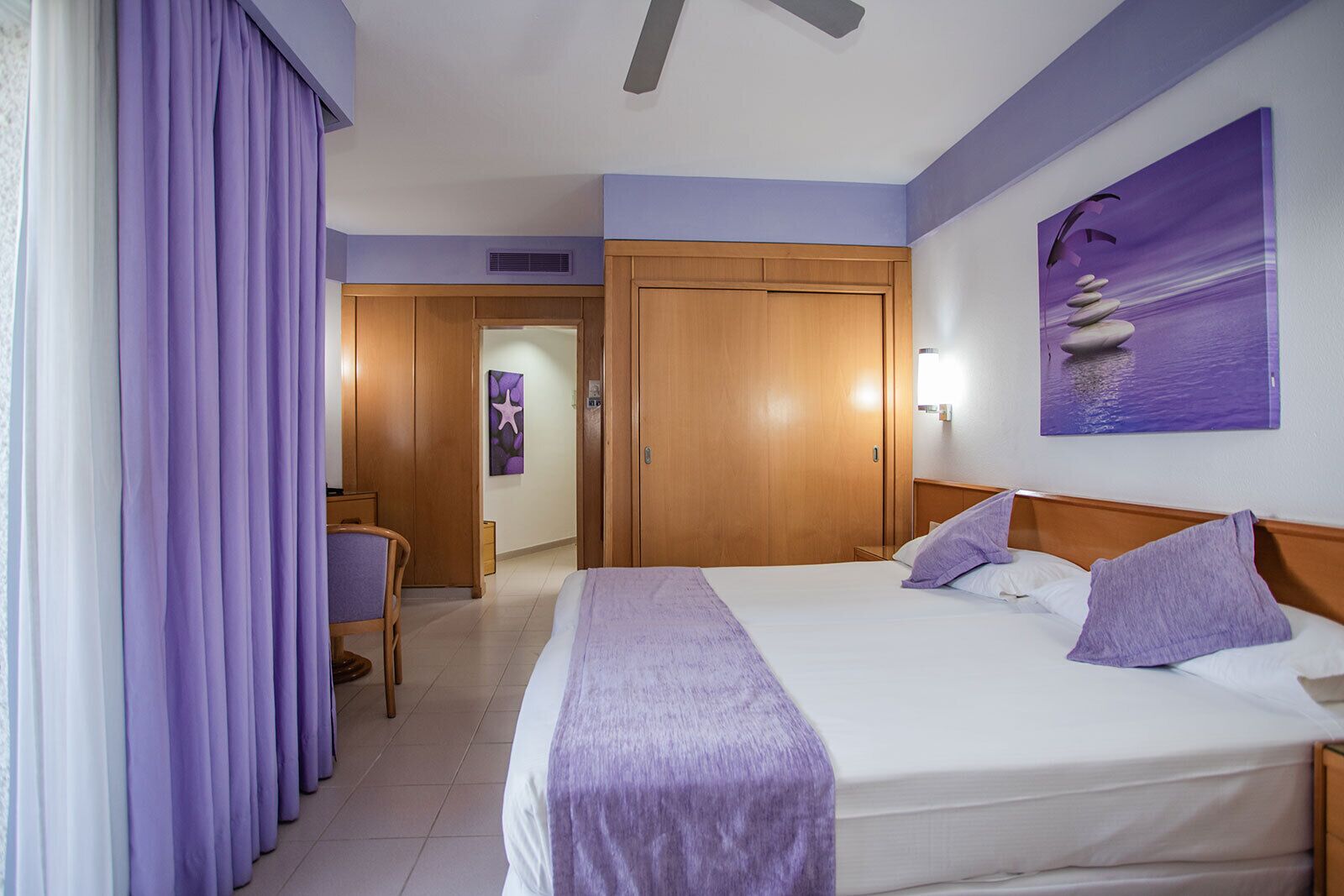 Een hotelkamer met een bed, een bureau en een stoel. Aan de muur hangt een schilderij en er is een raam met gordijnen.
