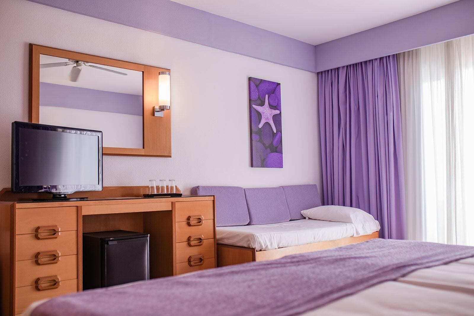A hotel room with a wooden desk, a television, a bed with a purple blanket, a nightstand, a mirror, and a starfish artwork.