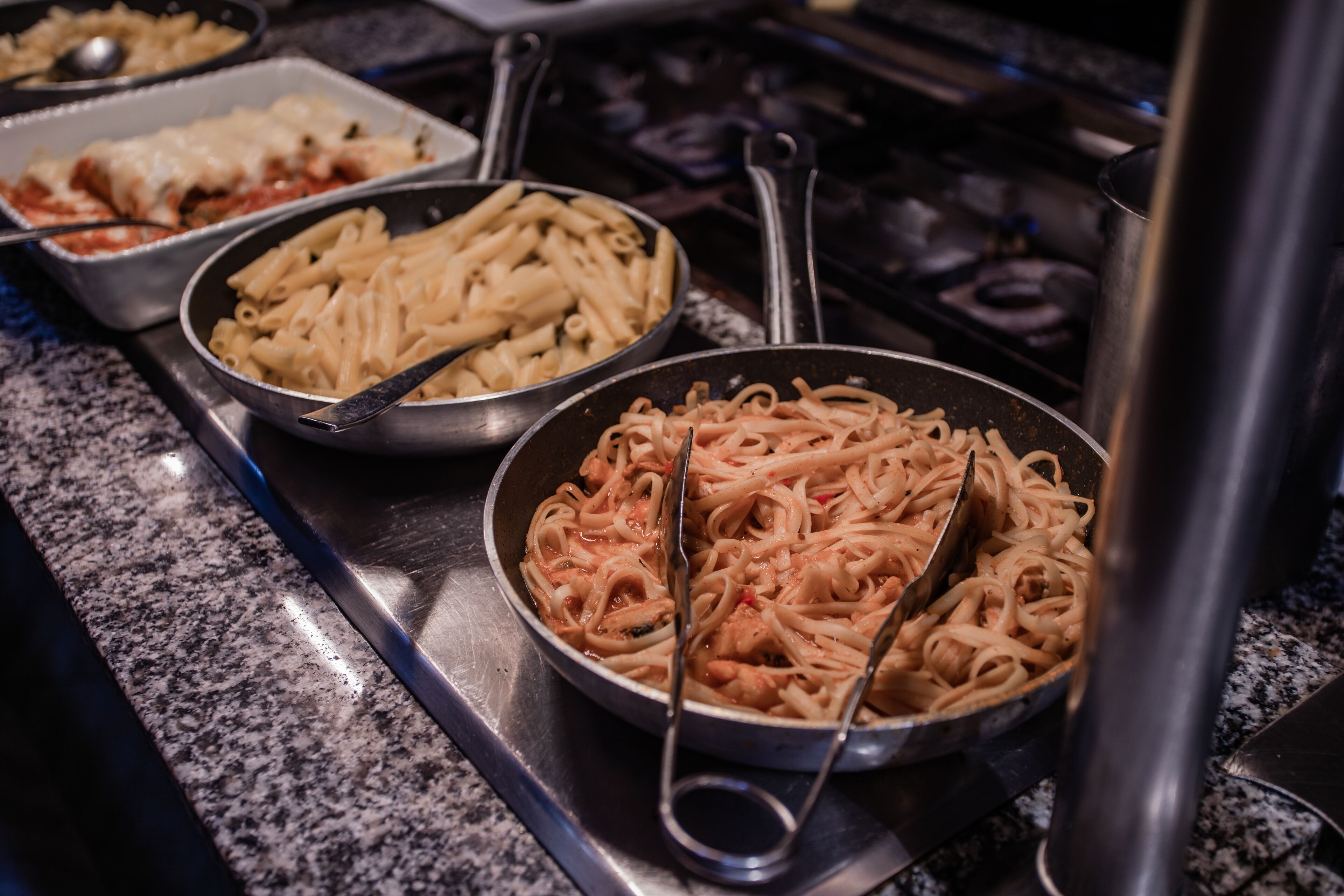 A buffet with pasta dishes, including penne and spaghetti, on a granite countertop.
