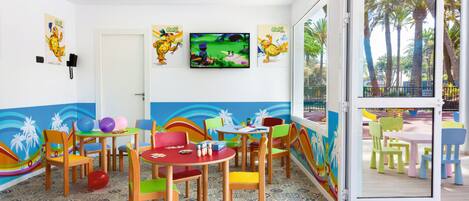 Un área de juegos infantiles con sillas y mesas coloridas, un televisor montado en la pared y un mural que representa una playa.