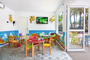 Un área de juegos infantiles con sillas y mesas coloridas, un televisor montado en la pared y un mural que representa una playa.