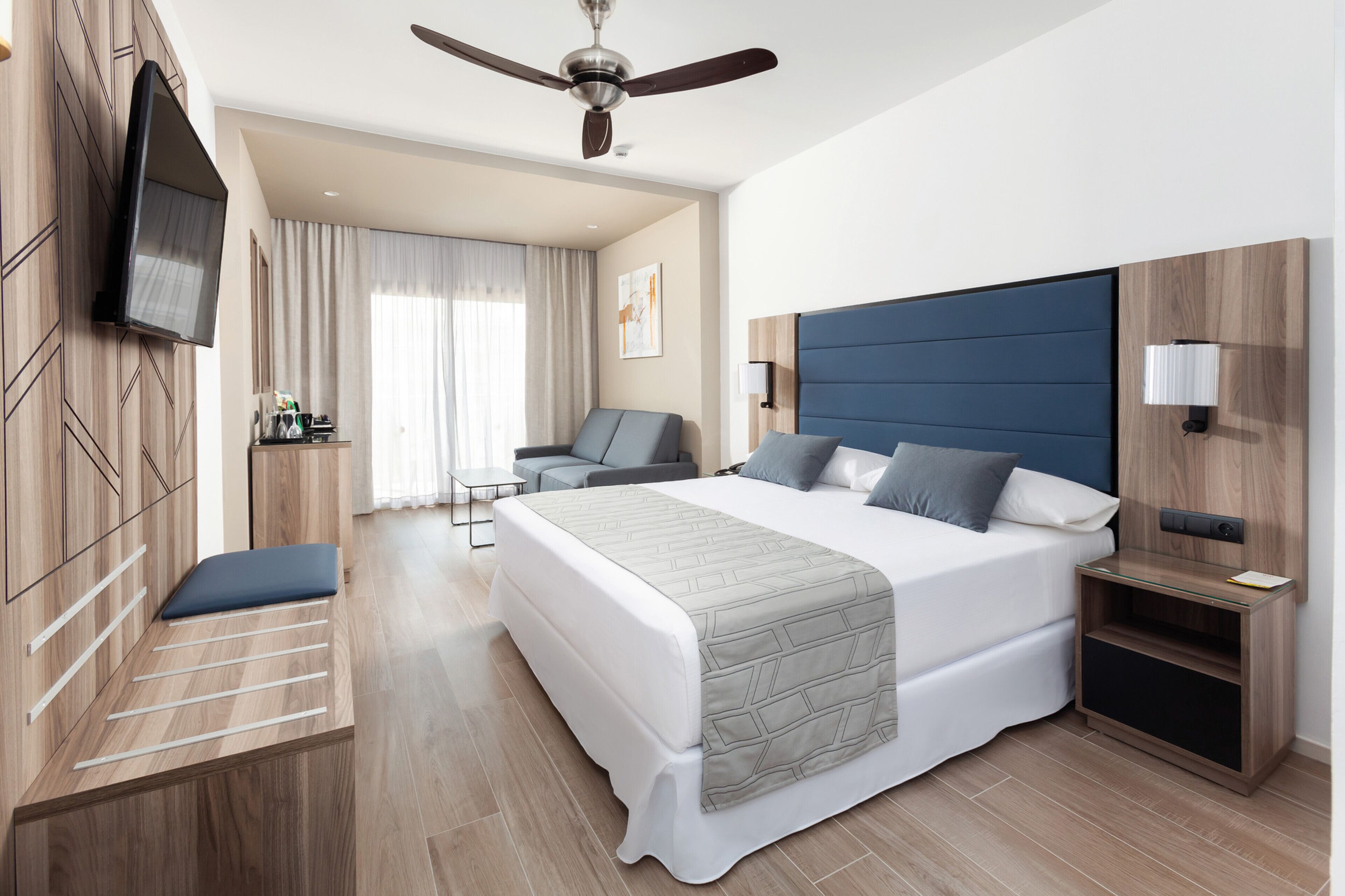 Habitación de hotel moderna con una cama grande, ventilador de techo, televisor de pantalla plana, escritorio y zona de estar.