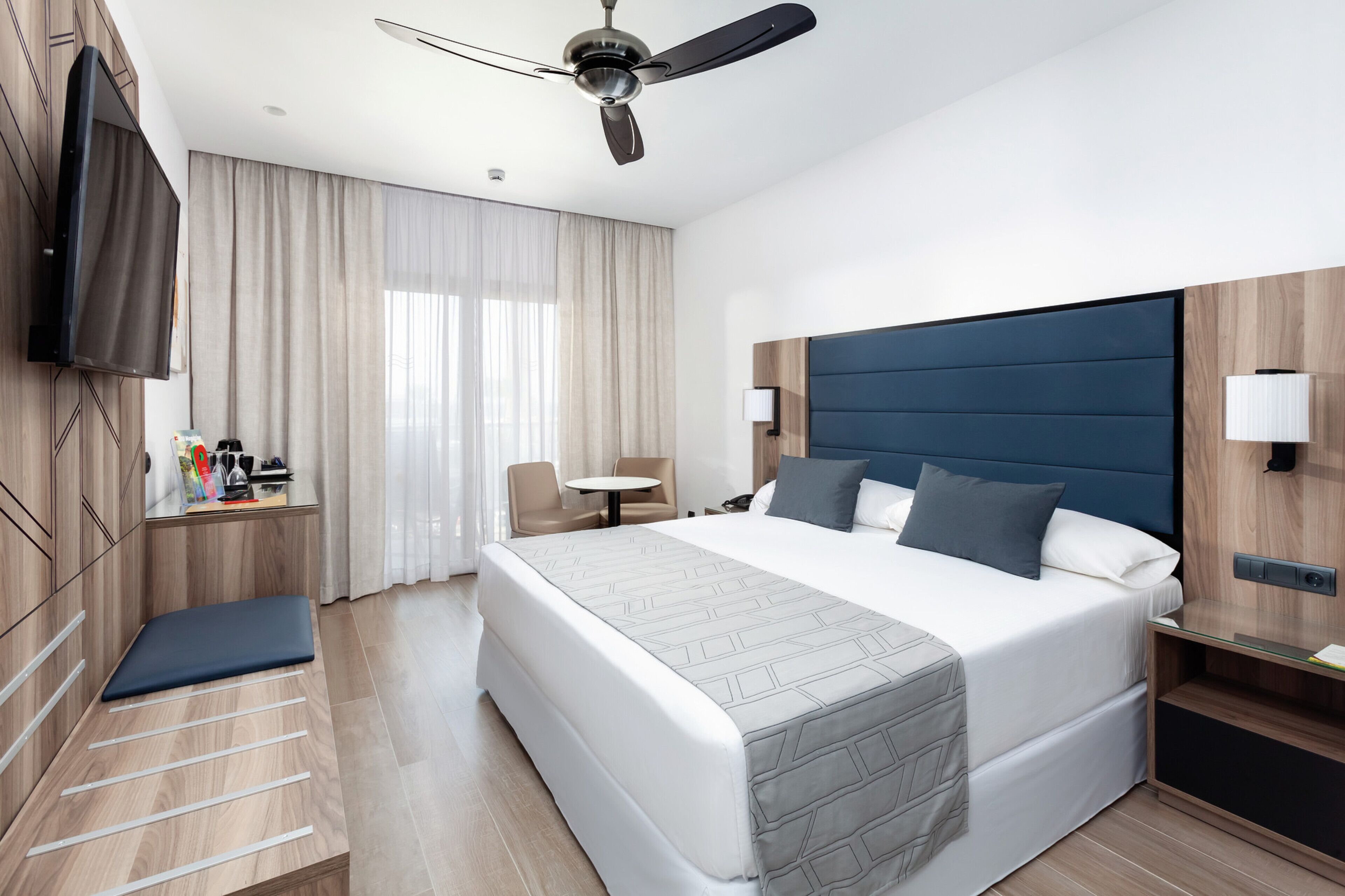 Une chambre d’hôtel moderne avec un grand lit, un ventilateur de plafond, un téléviseur fixé au mur, une petite table avec des chaises et une table de chevet avec une lampe.