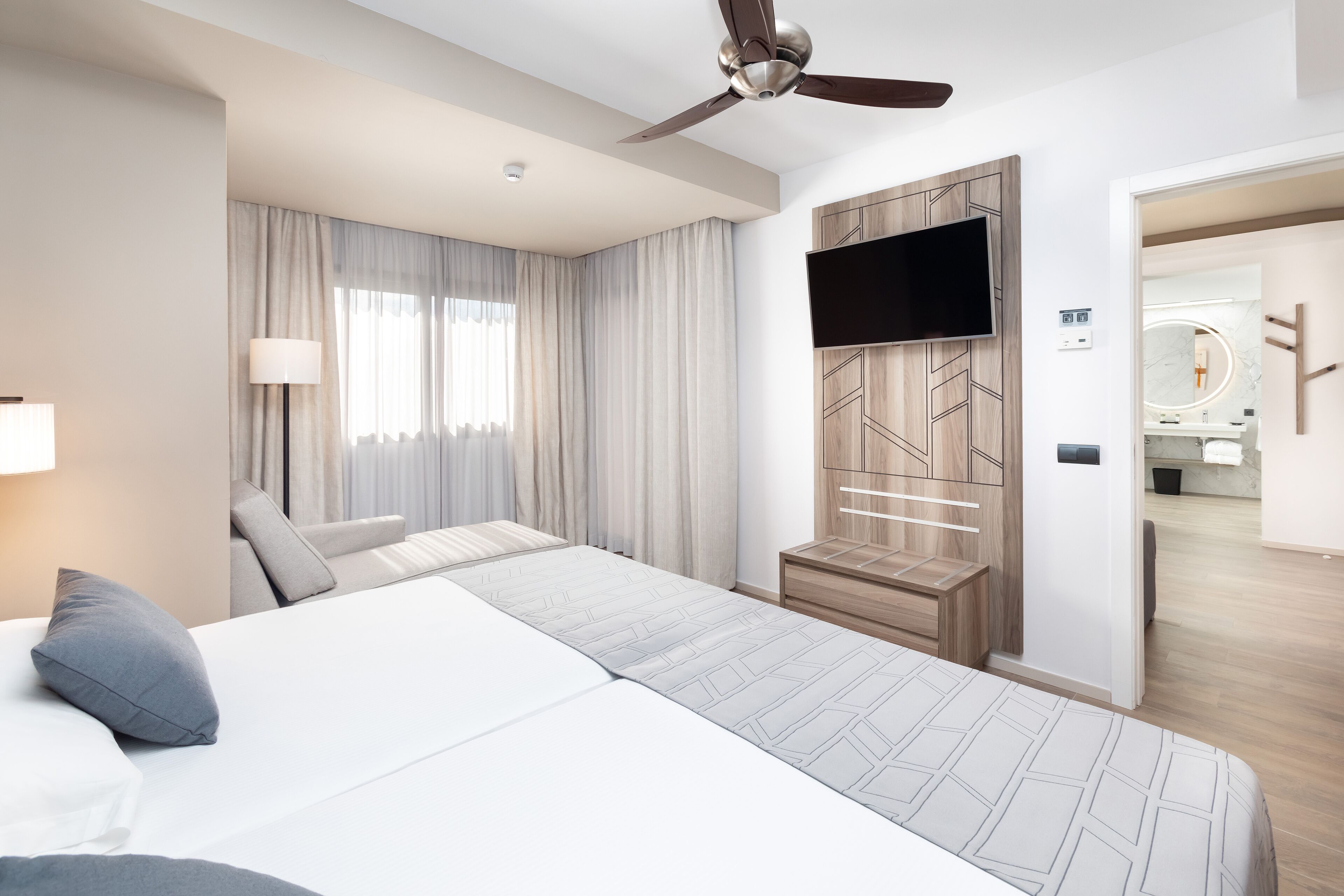 A modern hotel room with a large bed, a ceiling fan, a TV mounted on a wooden panel wall, and a bathroom visible through an open door.