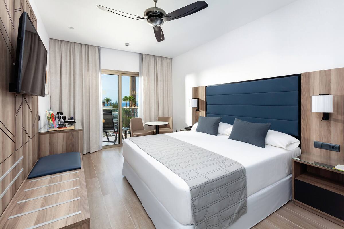 Een moderne hotelkamer met een groot bed, een plafondventilator, een flatscreen televisie, een bureau met lamp en uitzicht op het omliggende landschap.