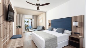 Une chambre d’hôtel dotée d’un grand lit, d’un ventilateur de plafond, d’un téléviseur fixé au mur, d’un coin salon avec une chaise et une table, et offrant une vue sur l’océan par la fenêtre.