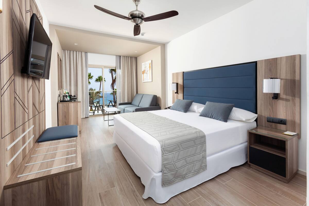 Hotelkamer met een groot bed, een plafondventilator, een aan de muur bevestigde televisie, een zithoek met een stoel en tafel, en uitzicht op de oceaan door het raam.