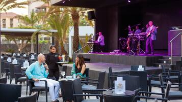 An outdoor dining area with wicker chairs and tables, a stage with musicians, and a palm tree in the background.