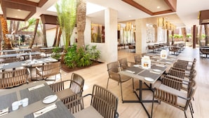 Un vaste espace intérieur aménagé en salle à manger, avec de nombreuses tables et chaises, un arrangement floral central et un plafond haut orné de poutres en bois.
