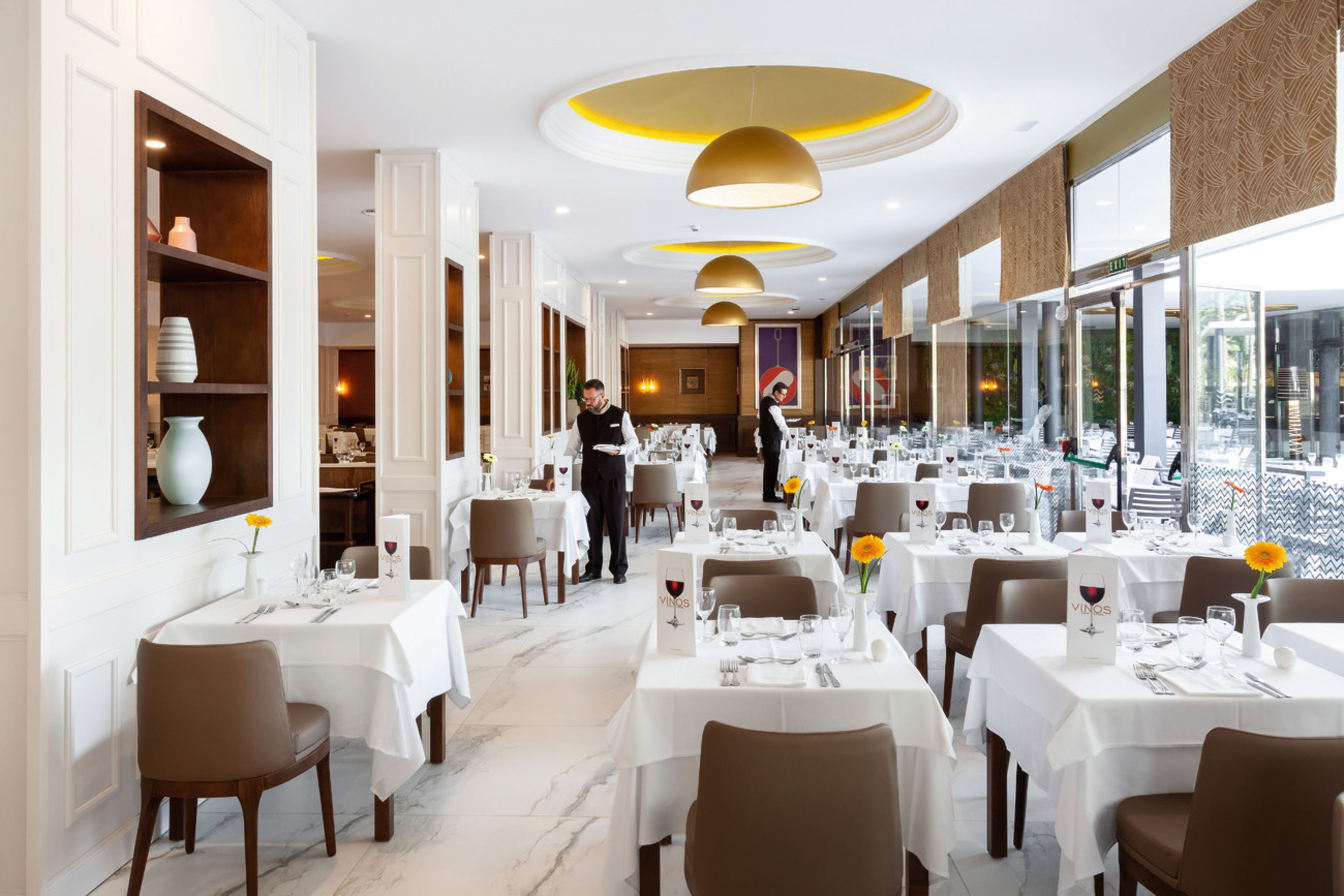 A restaurant with white tables, brown chairs, and a waiter.