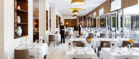 A restaurant with white tables, brown chairs, and a waiter.