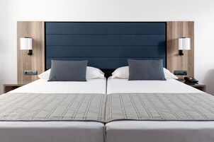 Ein Doppelbett mit dunklem Kopfteil, zwei Nachttischlampen und einer neutralen Bettdecke.