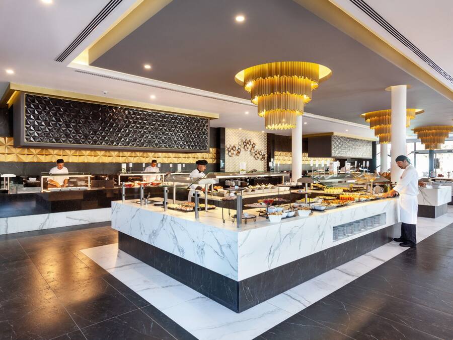 A modern hotel lobby with marble flooring, a central buffet area, and staff serving guests.