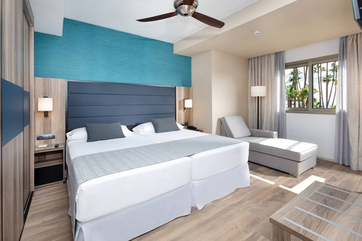 Een moderne hotelkamer met een groot bed, een bank, een bureau en een raam met uitzicht op palmbomen.