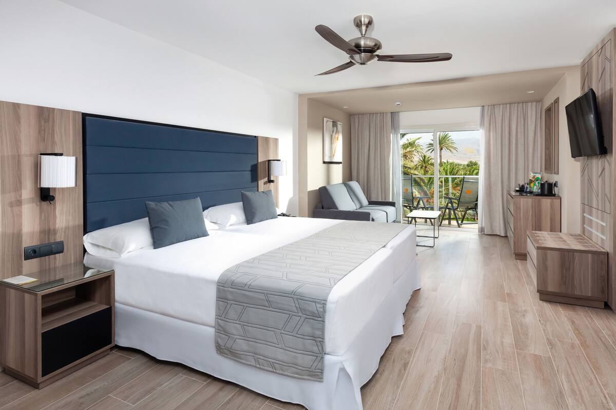 Een moderne hotelkamer met een groot bed, een bureau, een stoel, een televisie en een balkon met uitzicht op palmbomen.