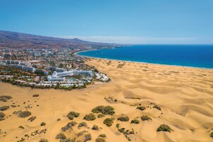 Vista aérea de un complejo turístico costero con playa de arena, un gran hotel y una cadena montañosa al fondo.
