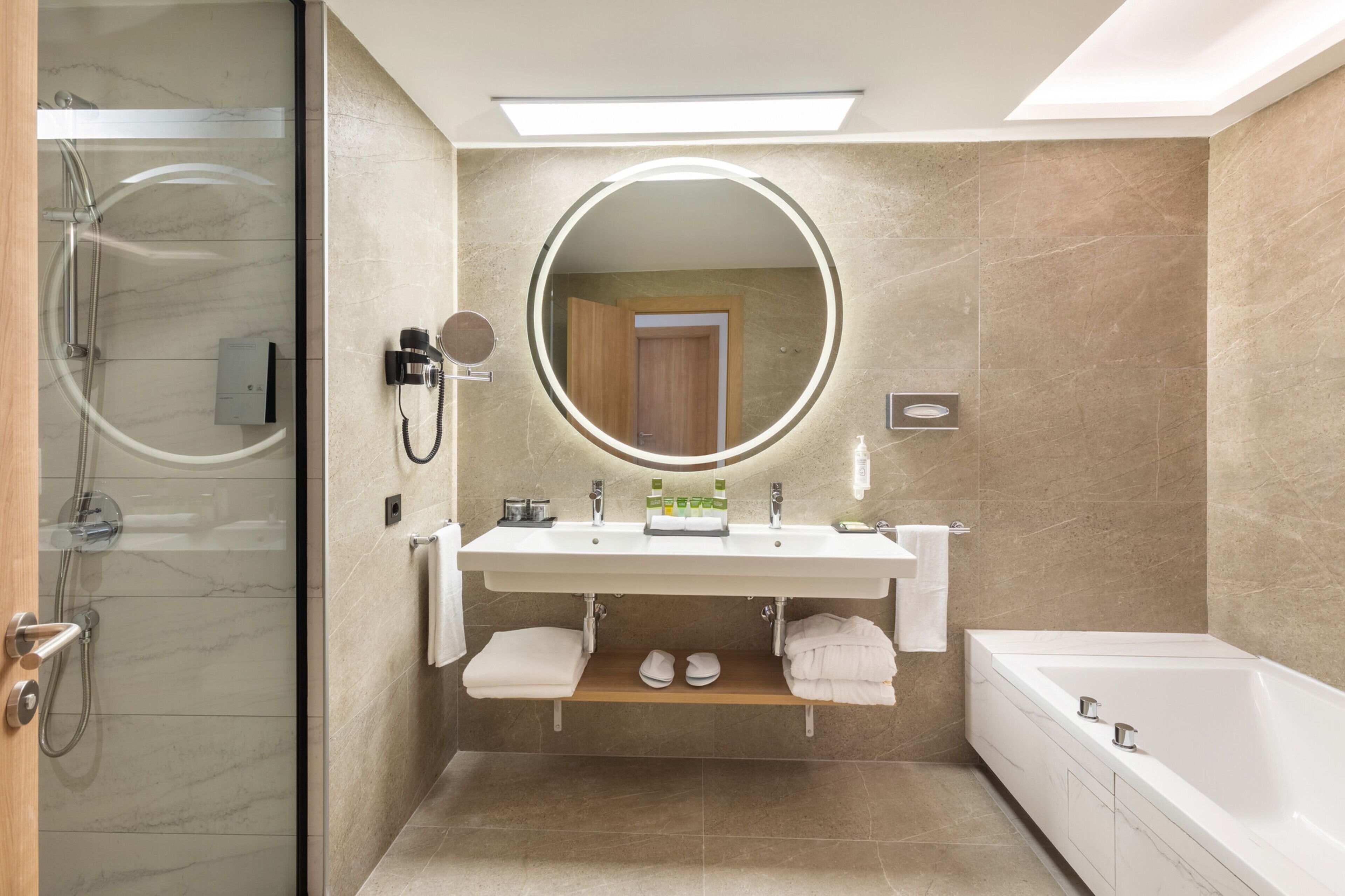Een moderne badkamer met een douche in een glazen cabine, een ronde spiegel, een witte wastafel en een bad.