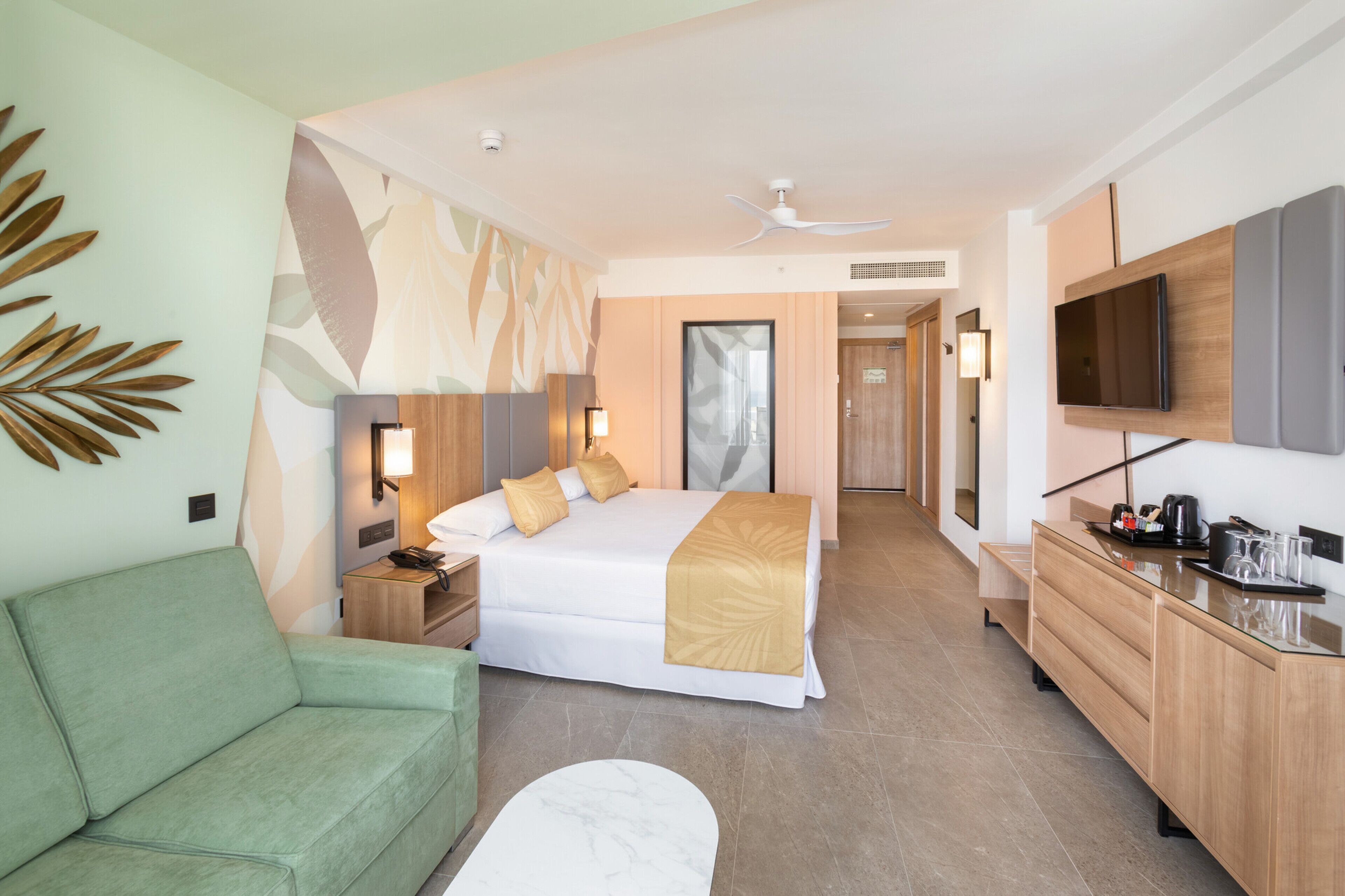Quarto de hotel com cama, sofá verde, escrivaninha com televisão e banheiro visível por uma porta aberta.
