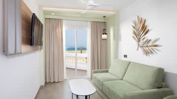 A modern living room with a green sofa, a flat-screen TV, a ceiling fan, and a view of the ocean through a sliding glass door.
