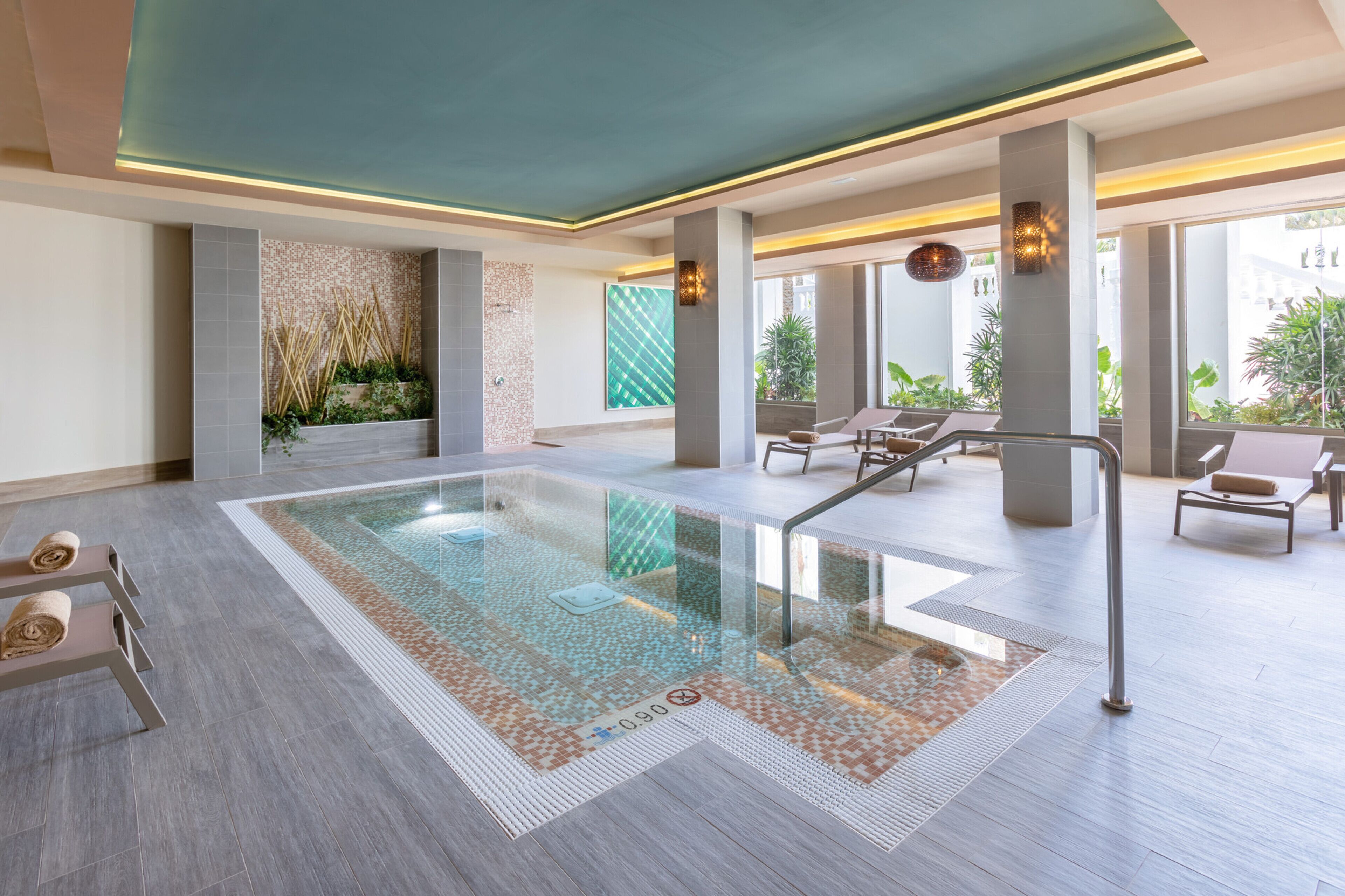 A modern indoor pool area with clear glass walls, a tiled floor, and a seating area with lounge chairs and a small table.
