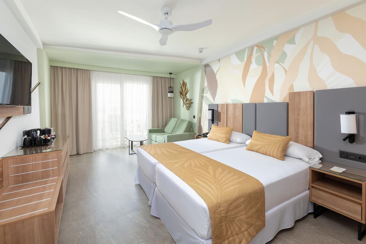 A hotel room with a large bed, a desk, a chair, a TV, and a ceiling fan.