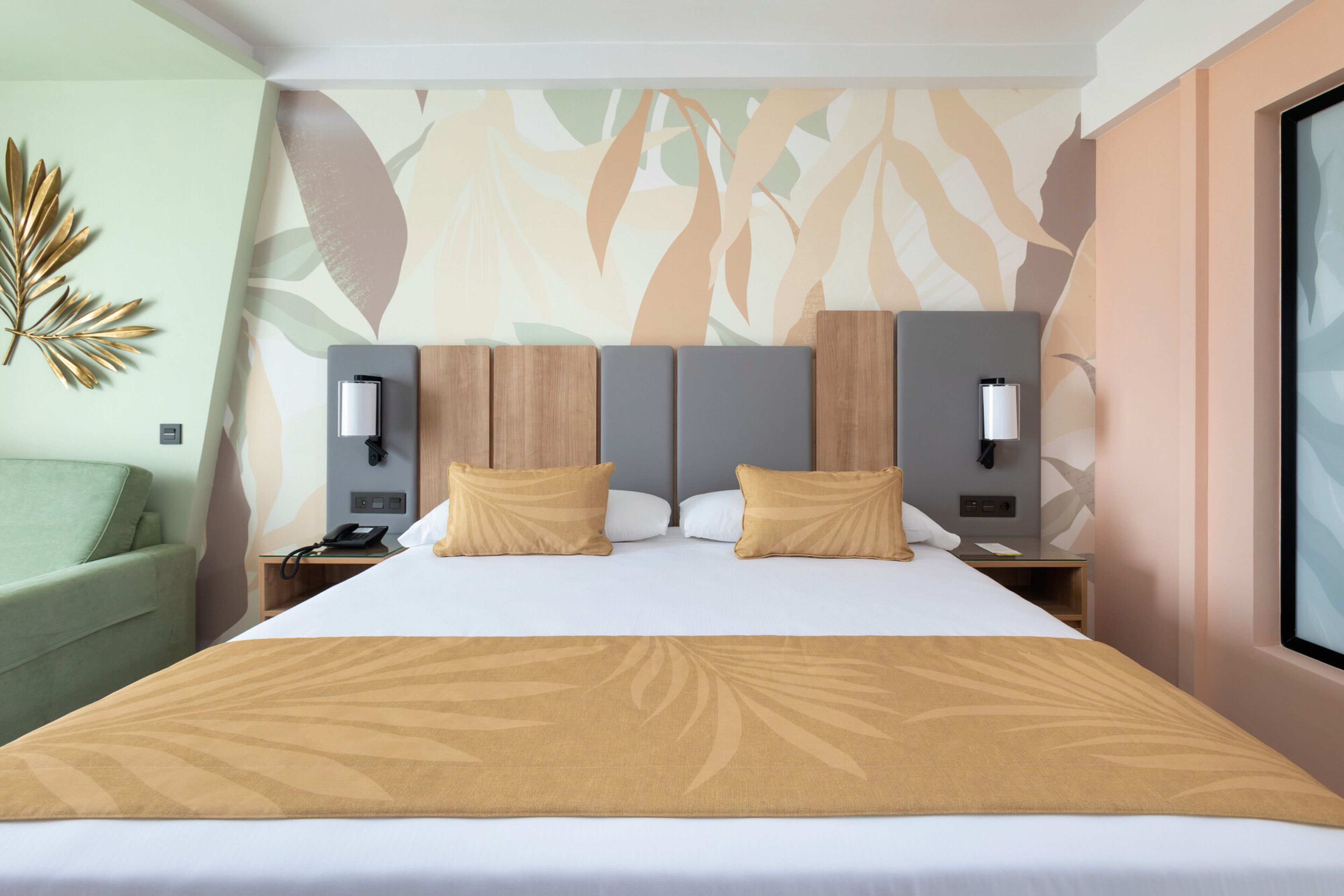 Een hotelkamer met een groot bed, twee nachtkastjes met lampen, een groene fauteuil, en een muur met bladmotief.
