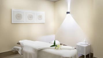 Un cuarto de spa con una camilla de masaje, una obra de arte colgada en la pared, una lámpara blanca y una pequeña mesita auxiliar blanca con una botella.