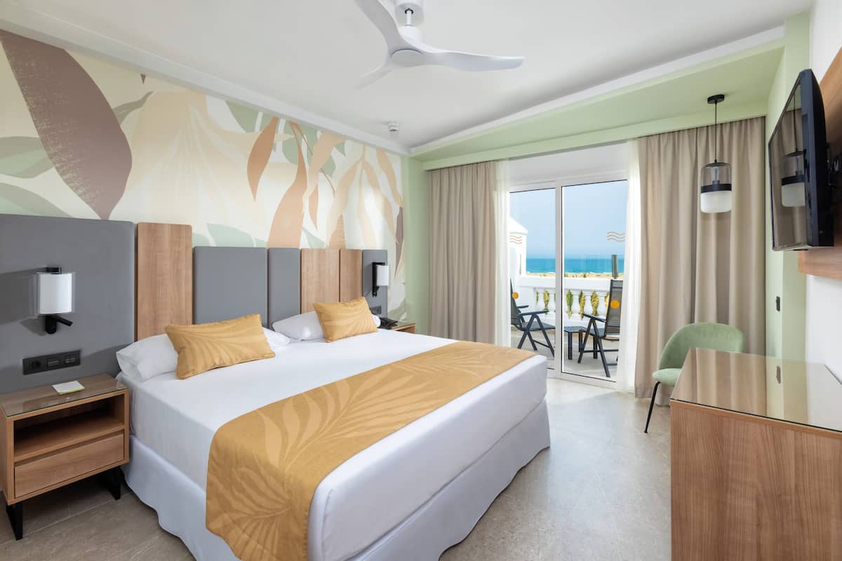 A hotel room with a large bed, a desk, a chair, a television, and a balcony with a view of the beach.