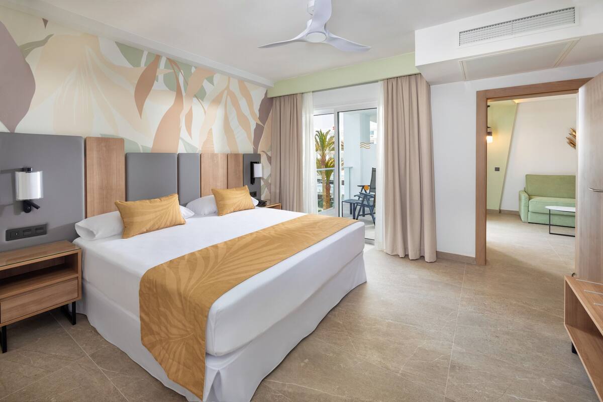 A modern hotel room with a large bed, a ceiling fan, a nightstand, and a view of the balcony.