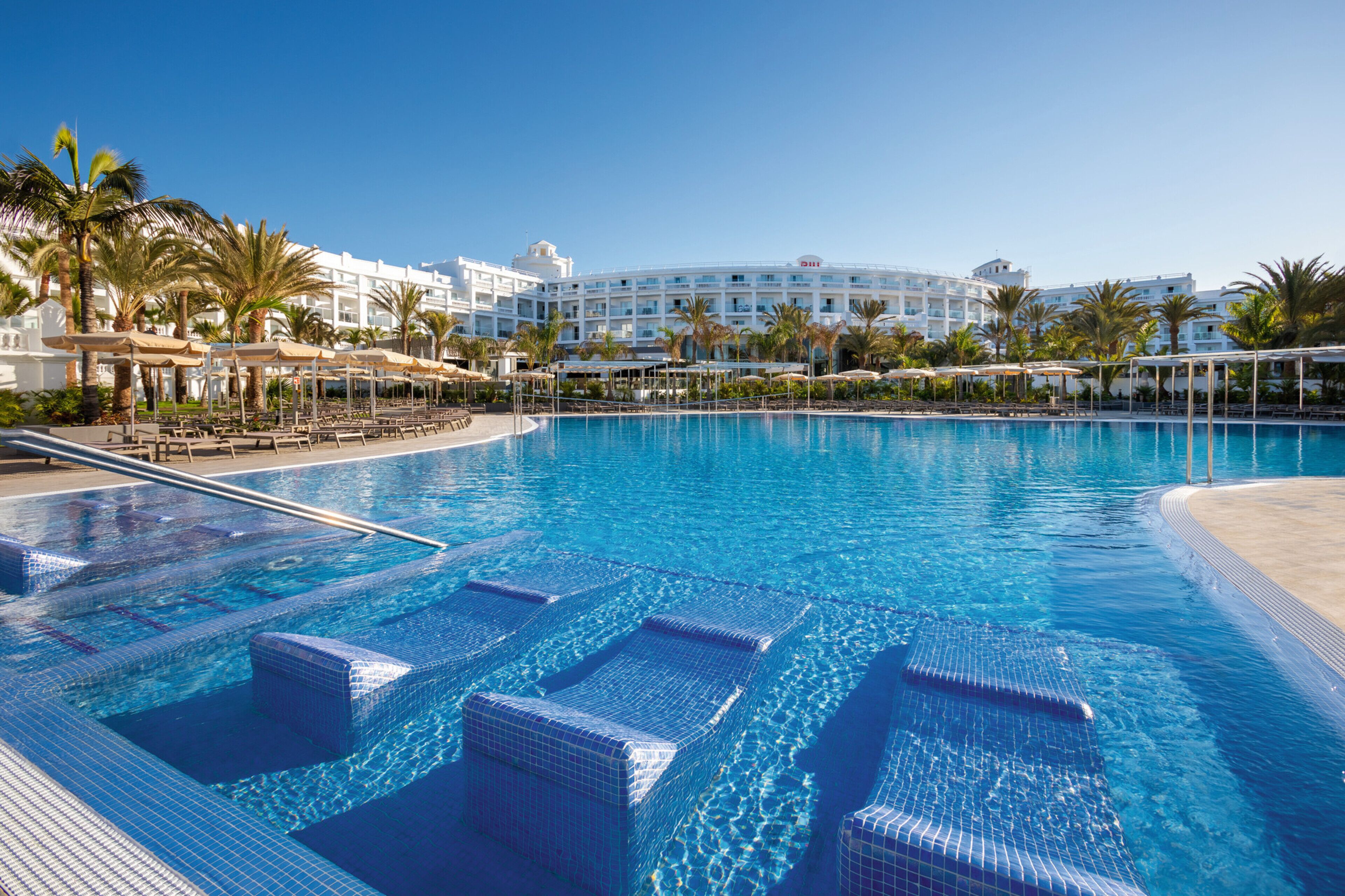 A large, clear swimming pool with lounge chairs and umbrellas, surrounded by palm trees and a hotel building in the background.
