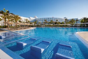 A large, clear swimming pool with lounge chairs and umbrellas, surrounded by palm trees and a hotel building in the background.
