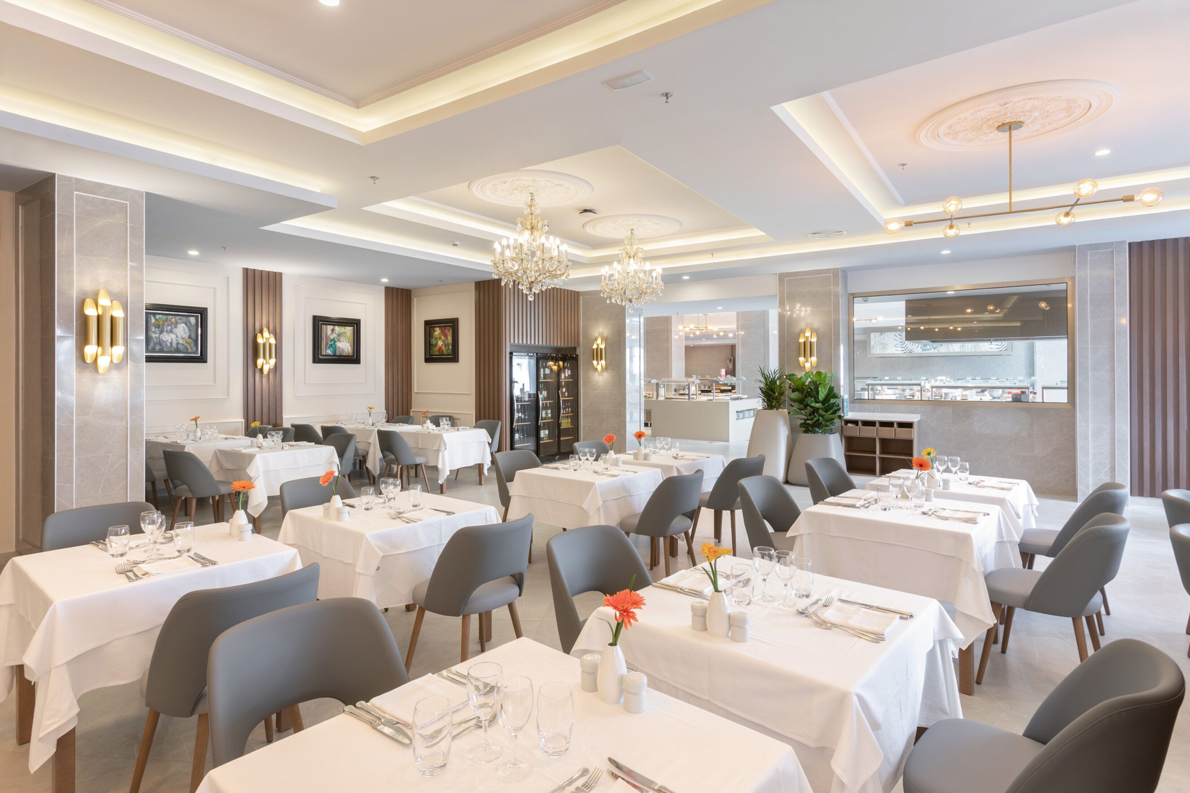 A dining area with multiple tables set for service, a chandelier, and a view of the kitchen through a glass partition.