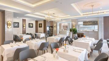 A dining area with multiple tables set for service, a chandelier, and a view of the kitchen through a glass partition.