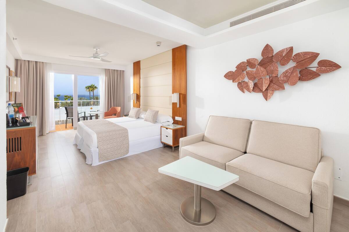 A hotel room with a bed, a sofa, a small table, a nightstand, and a wall-mounted decorative piece.