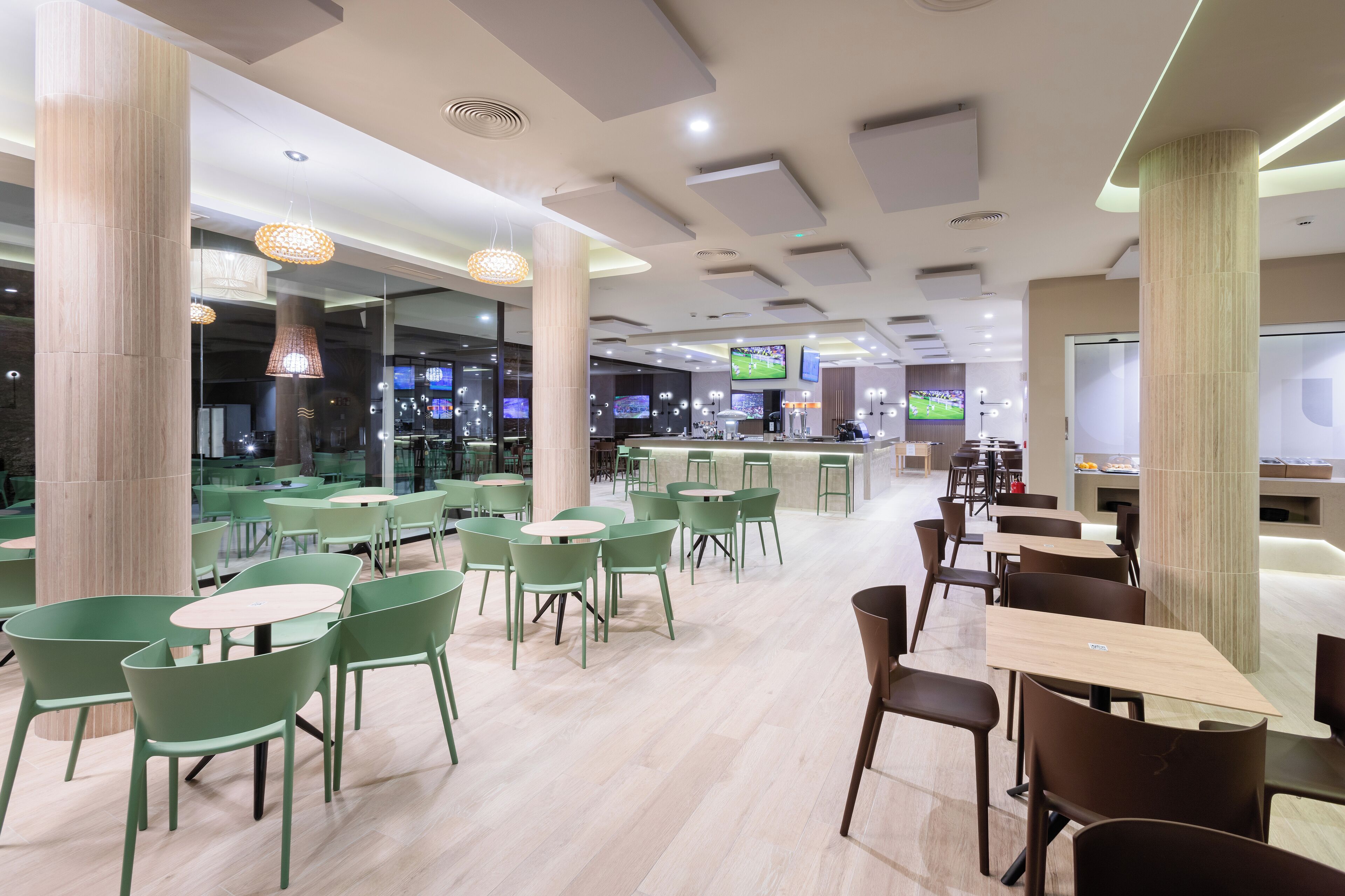 Ein modernes Restaurant mit grünen und braunen Stühlen, Holztischen und einer Bar mit Bildschirmen, die Sportübertragungen zeigen.