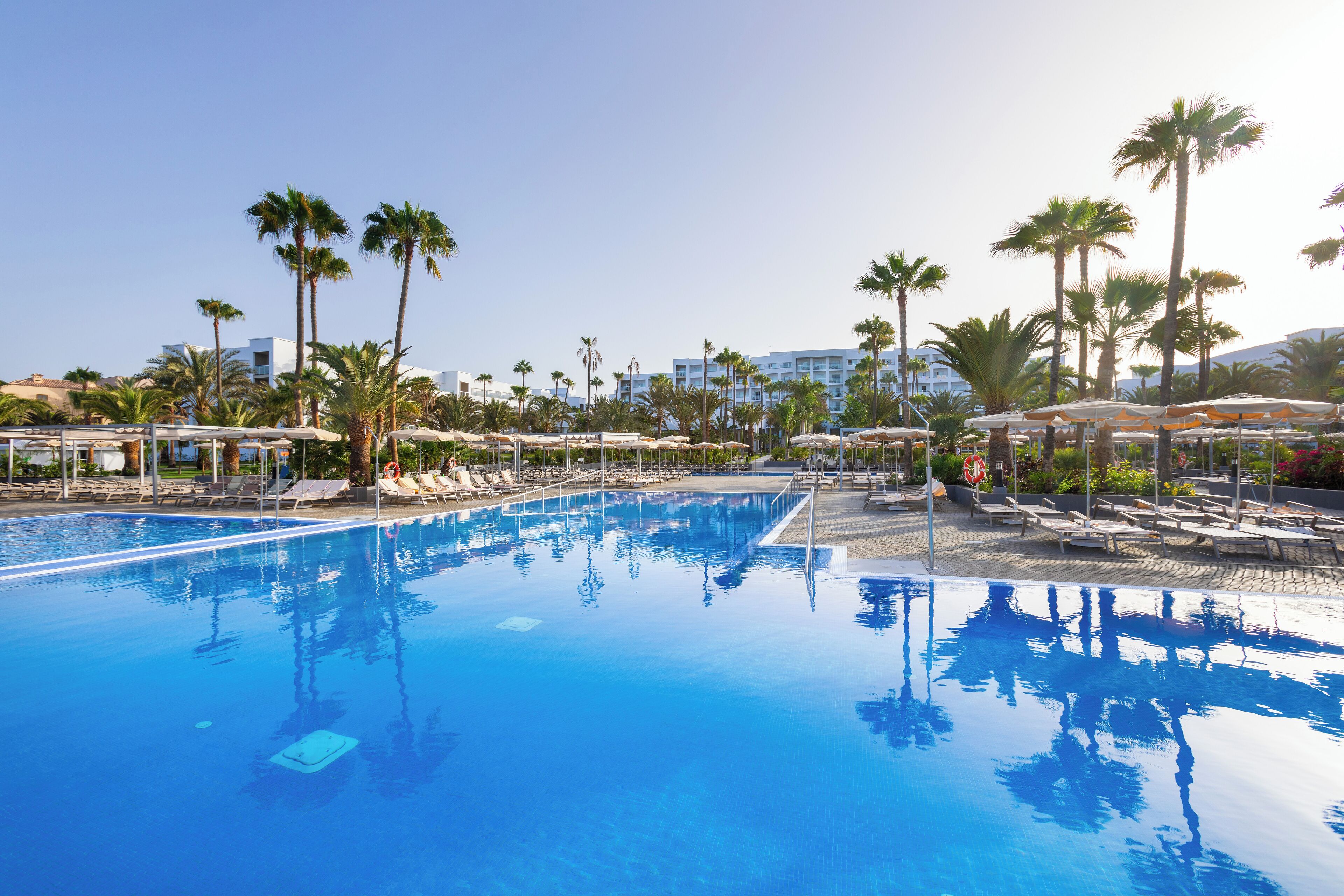 A large swimming pool with sun loungers and palm trees, surrounded by a hotel building.