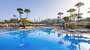 A large swimming pool with sun loungers and palm trees, surrounded by a hotel building.