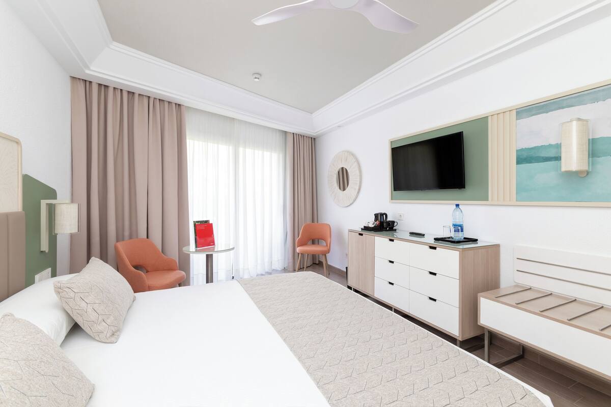 A hotel room with a large bed, a desk with a chair, a television, and a window with curtains.