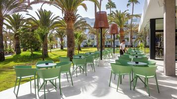 An outdoor patio with green chairs and tables, surrounded by palm trees and a building in the background.