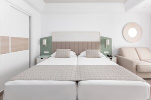 Una cama doble con cubrecama acolchado, dos mesitas de noche con lámparas, un espejo redondo en la pared y un sofá beige.