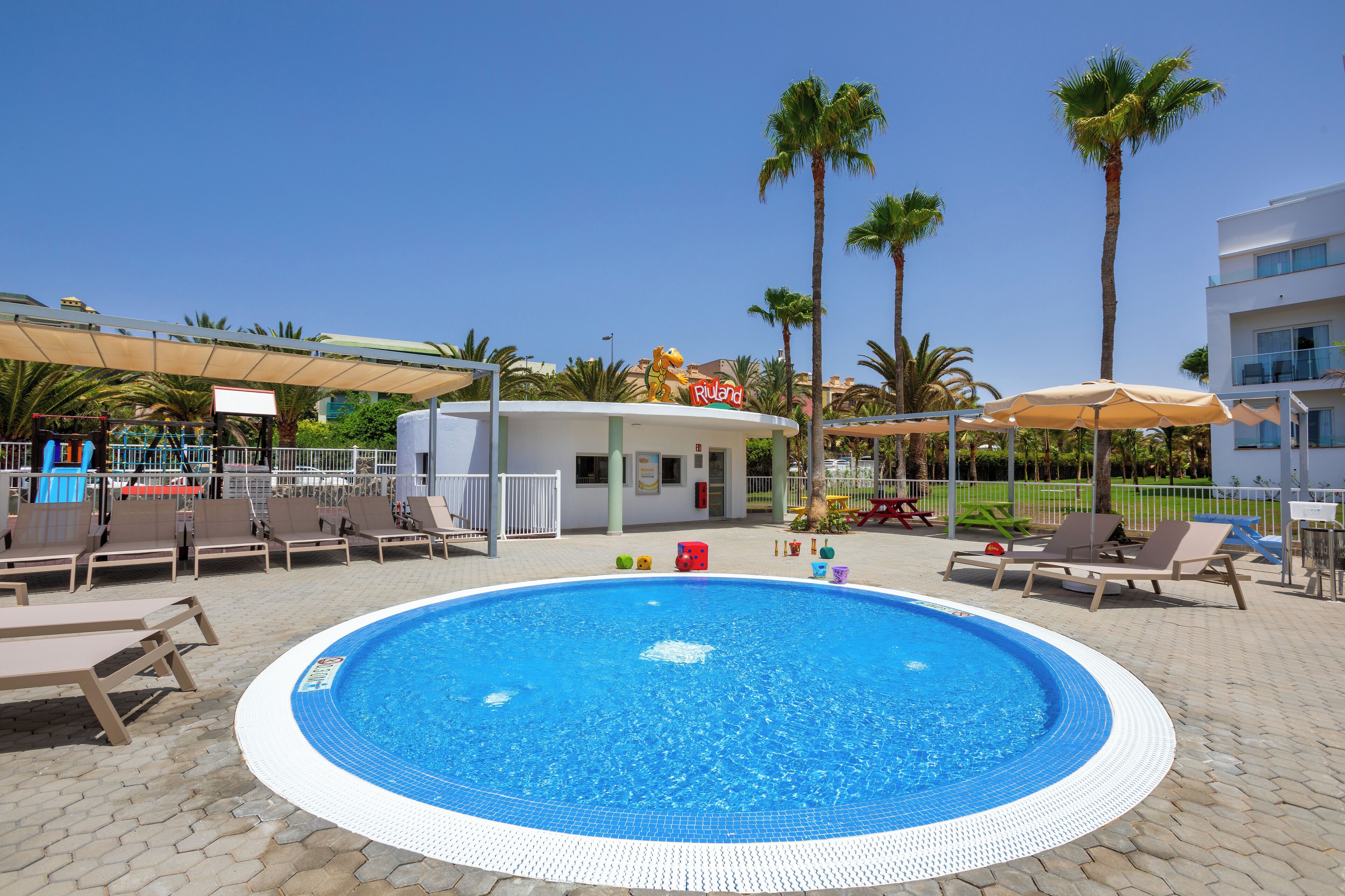 A swimming pool with lounge chairs and umbrellas, surrounded by palm trees and a building with a sign.