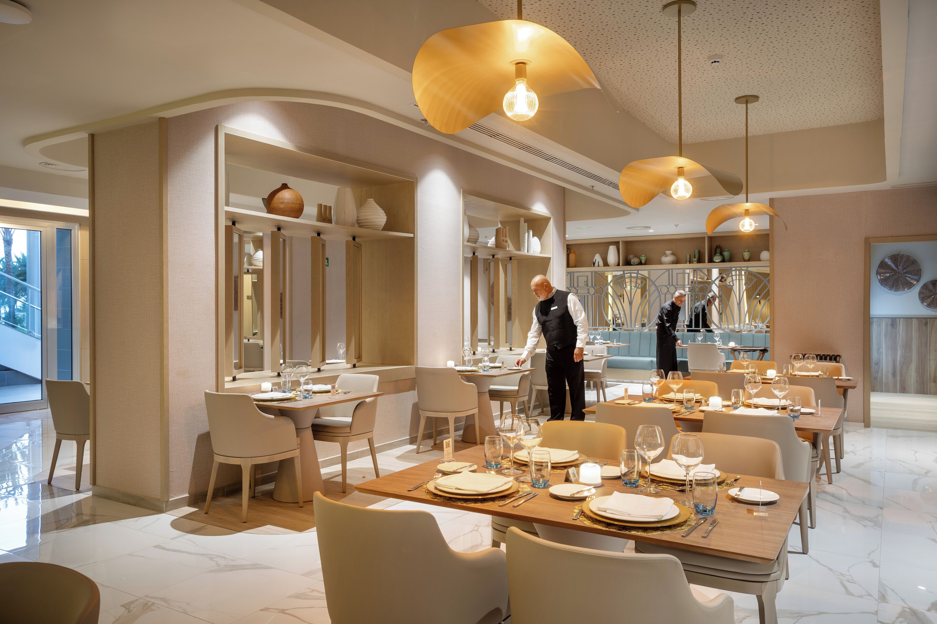 A modern dining area with tables set for service, a waiter attending to guests, and a view of the kitchen in the background.