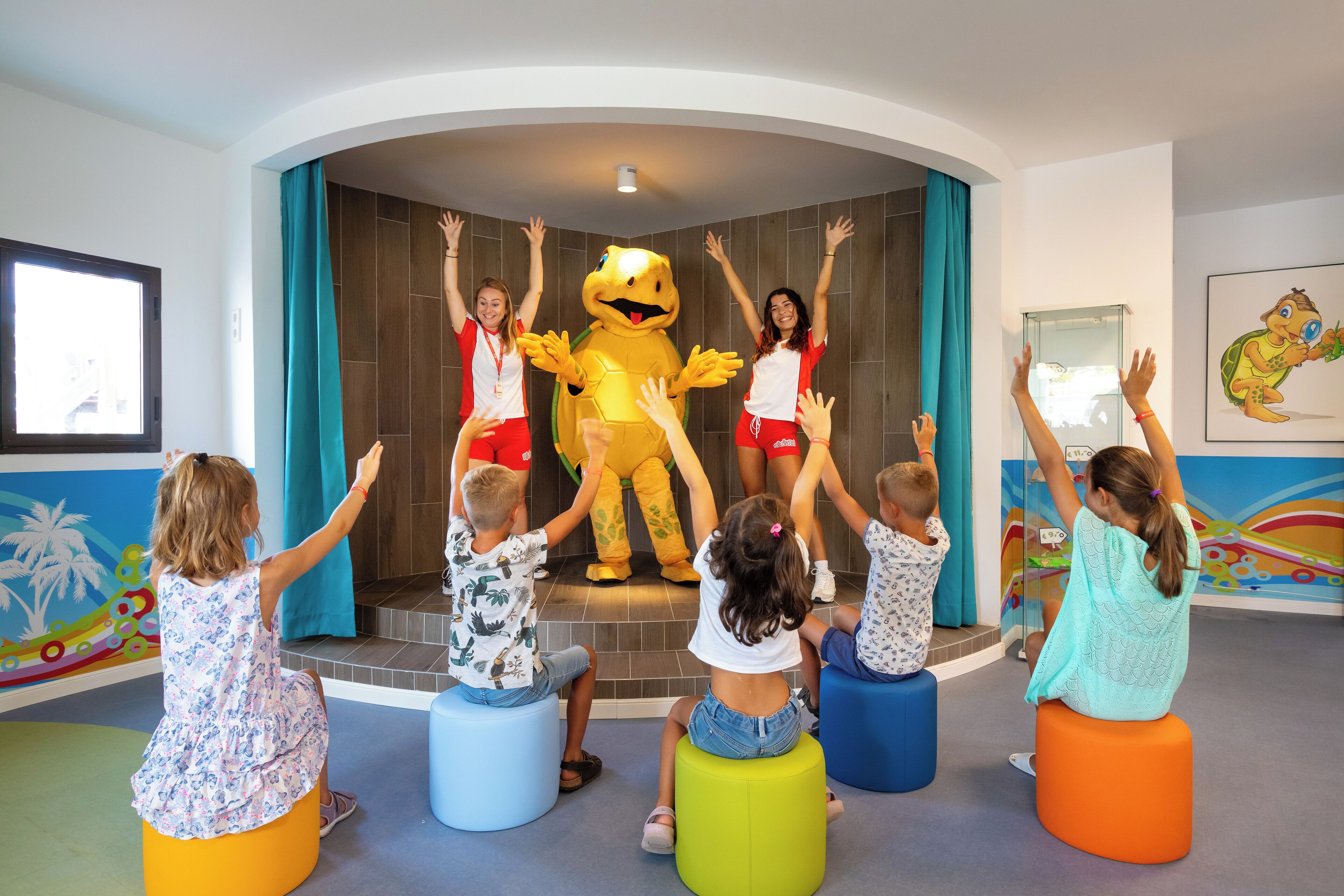 Children sitting on colorful stools facing a mascot in a room with a playful mural and a large window.
