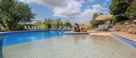 Una piscina cristalina con tumbonas y sombrillas, rodeada de vegetación y un muro de piedra.