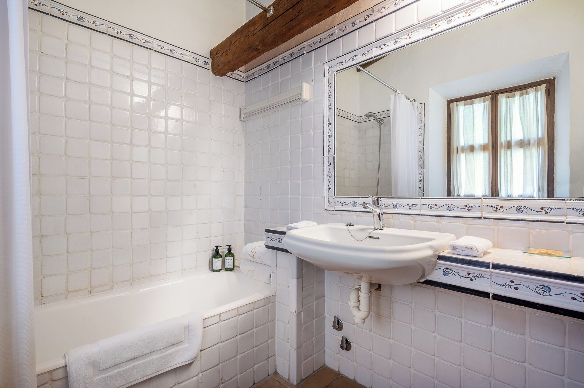 Ein Badezimmer mit weißen Fliesen, einer weißen Badewanne, einem Waschbecken mit Spiegel und einem Fenster mit Vorhängen.