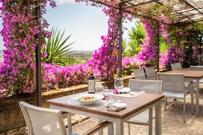 Zona d'exterior per dinar amb taules i cadires, envoltada de flors roses i vegetació exuberant.