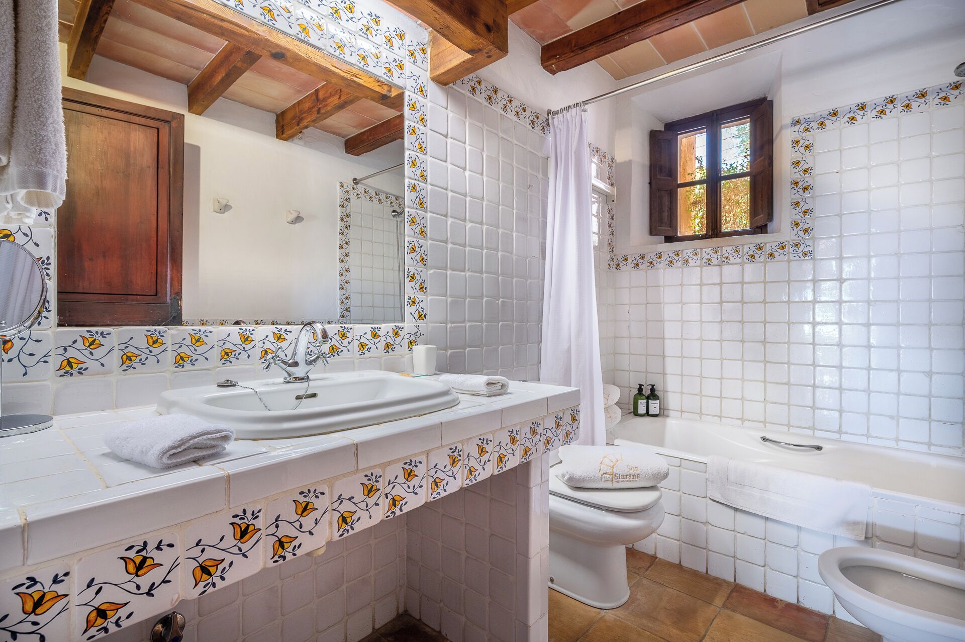 Casa de banho com lavatório, sanita e banheira, apresentando padrões de azulejos florais e vigas de madeira.